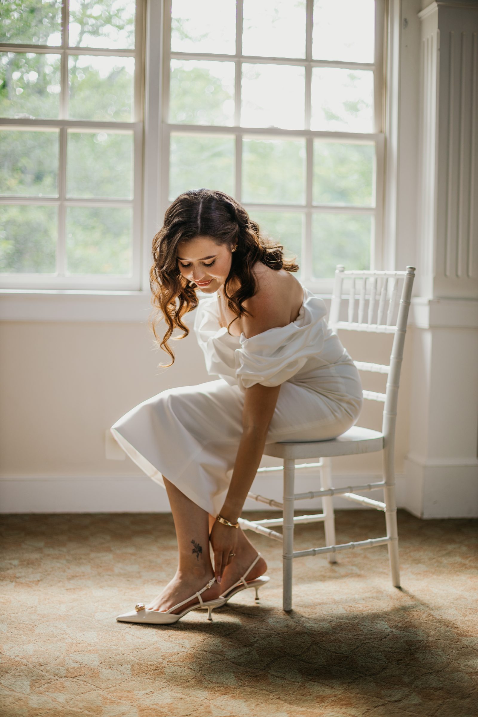Bride fastening her shoe by the window — candid bridal portrait